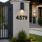 A close-up shows the address digits "4579" in a dark charcoal metal finish mounted on a modern concrete gate post. The scene includes a vertical-slat security gate to the left and a softly lit concrete walkway leading toward a luxury home in the background.