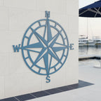 A large light blue metal compass rose sign fixed to an outdoor cream-colored tiled wall by a swimming pool, with a glimpse of a marina dock and boats in the background.