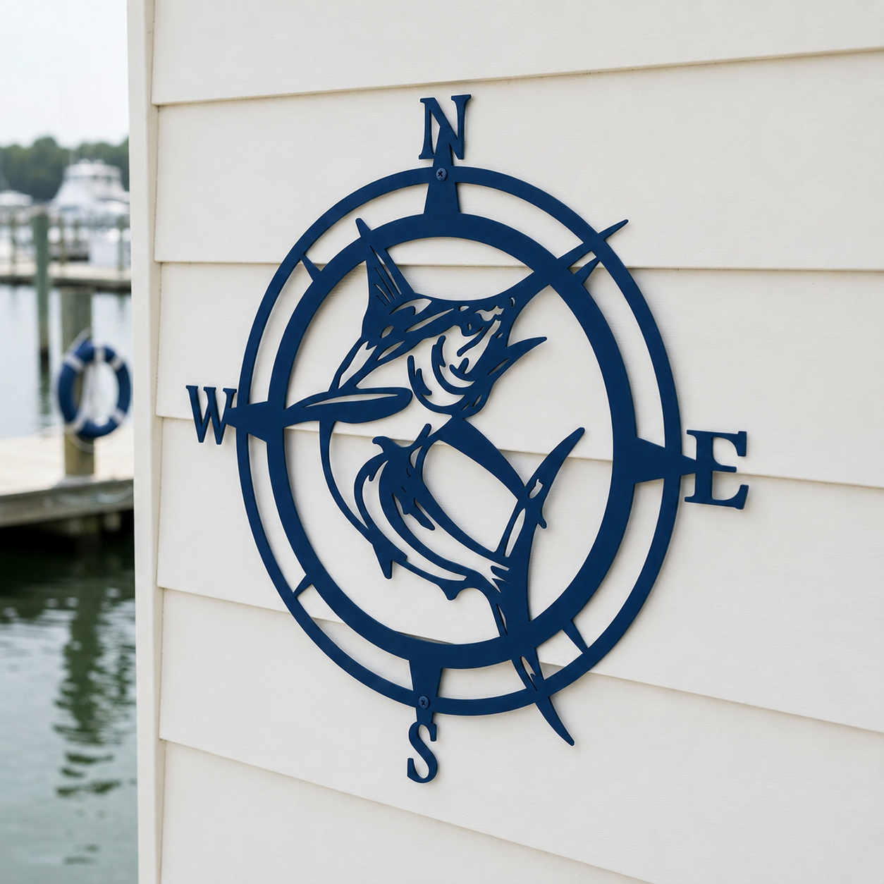 Navy blue finish of a metal marlin fish inside a compass frame, mounted on a white siding wall with a boat dock and lake visible in the background.