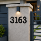 A close-up view of dark charcoal-colored house numbers "3163" installed on a concrete gate pillar. A warm light glows directly above the numbers, with a dark horizontal slat privacy fence on the left and a stone paver walkway visible behind a modern metal gate.