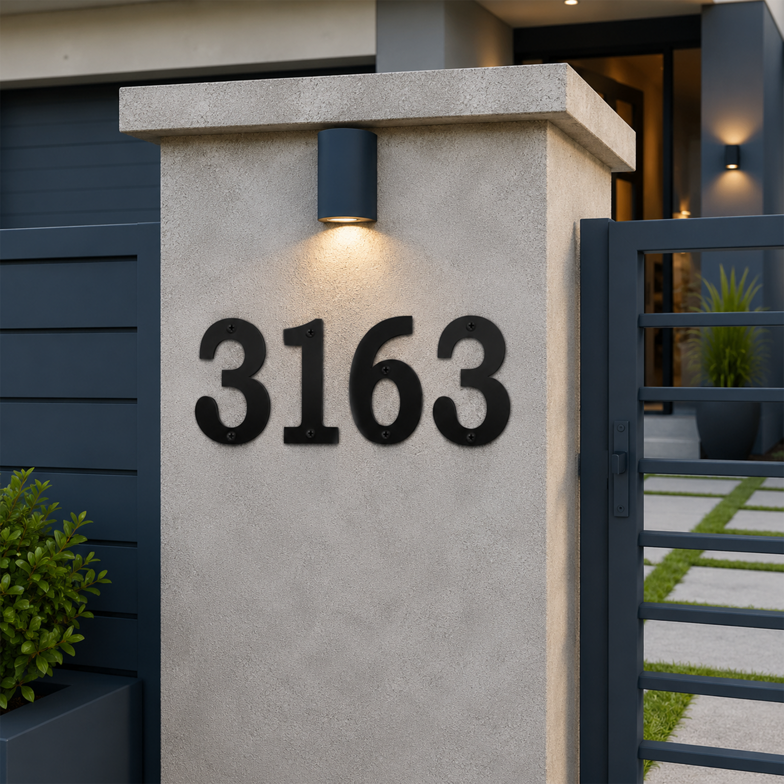 A close-up view of dark charcoal-colored house numbers "3163" installed on a concrete gate pillar. A warm light glows directly above the numbers, with a dark horizontal slat privacy fence on the left and a stone paver walkway visible behind a modern metal gate.