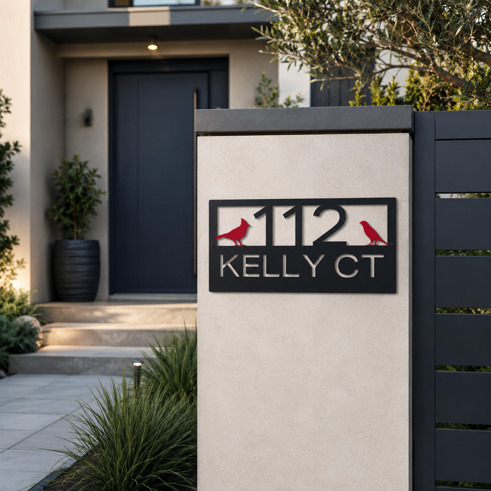 A charcoal-finished metal address sign with red cardinal accents is mounted to a smooth concrete gate pillar at a property entrance. The numbers and street name "112 KELLY CT" are clearly legible, set against a backdrop of a dark horizontal privacy fence and ornamental grasses.
