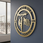 An elegant satin silver metallic circular sign featuring a detailed pine forest and mountain range cutout design with the name "THE MILLERS" on the upper rim. The plaque is installed on a modern taupe horizontal wood wall overlooking a sunlit deck space.