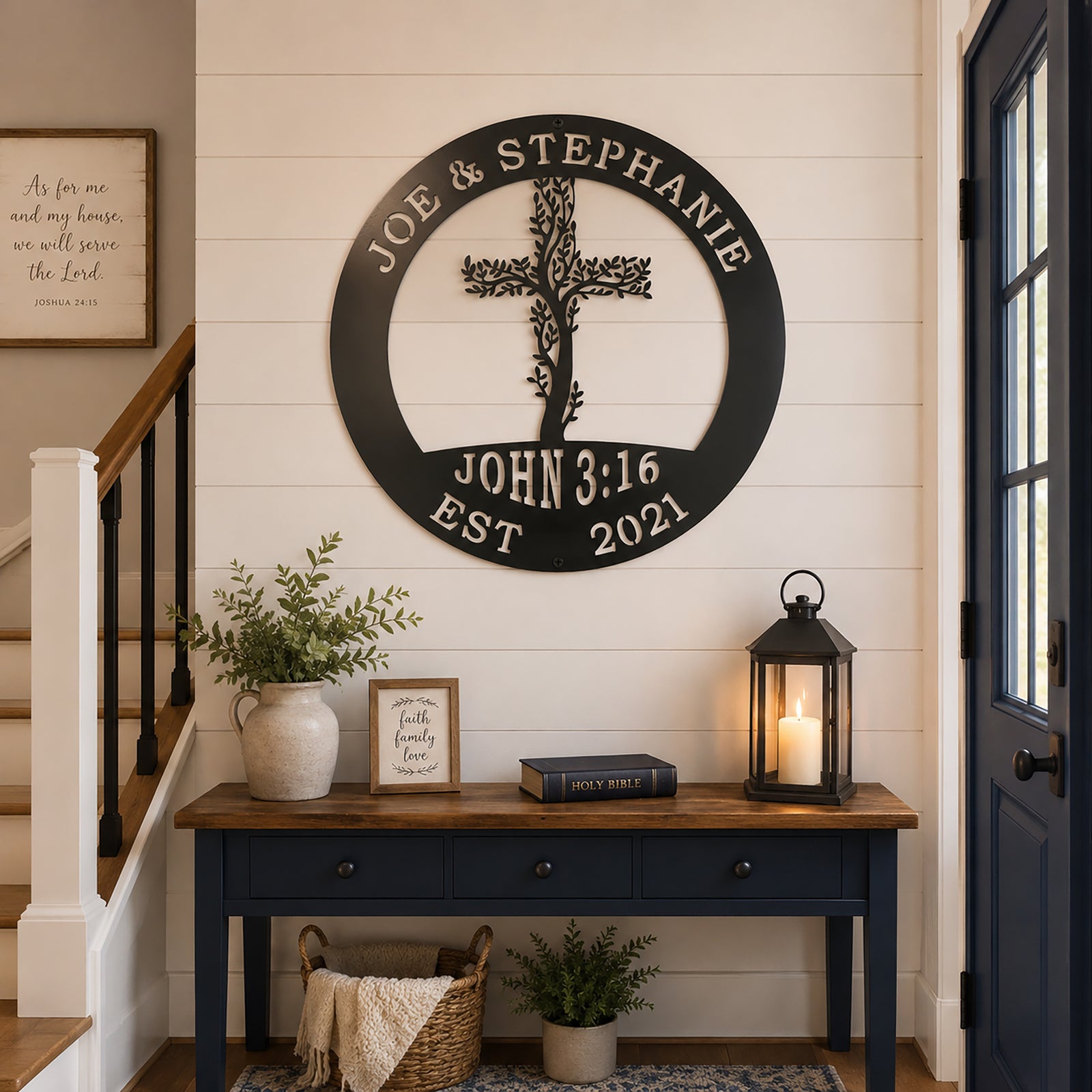 A textured satin black circular metal plaque reading "JOE & STEPHANIE EST 2021" and "JOHN 3:16" is centered on a white shiplap entryway wall. The sign is positioned directly above a dark blue console table arranged with a Holy Bible, a candle lantern, a ceramic pitcher, and framed scripture art.