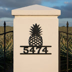 Black pineapple metal address plaque with house number 5474 displayed on a white entrance pillar by a metal gate