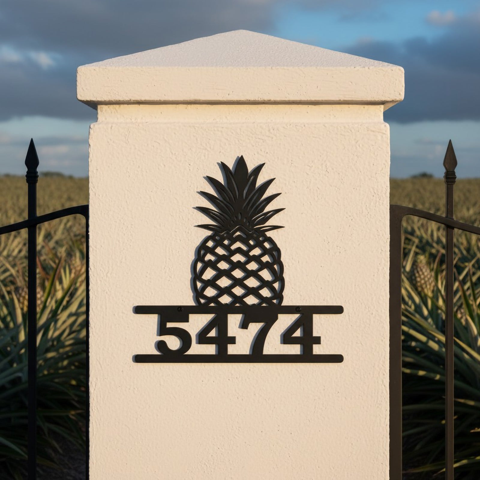 Black pineapple metal address plaque with house number 5474 displayed on a white entrance pillar by a metal gate