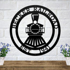 Black metal locomotive wall plaque with personalized name and year mounted on white brick with floral accents.