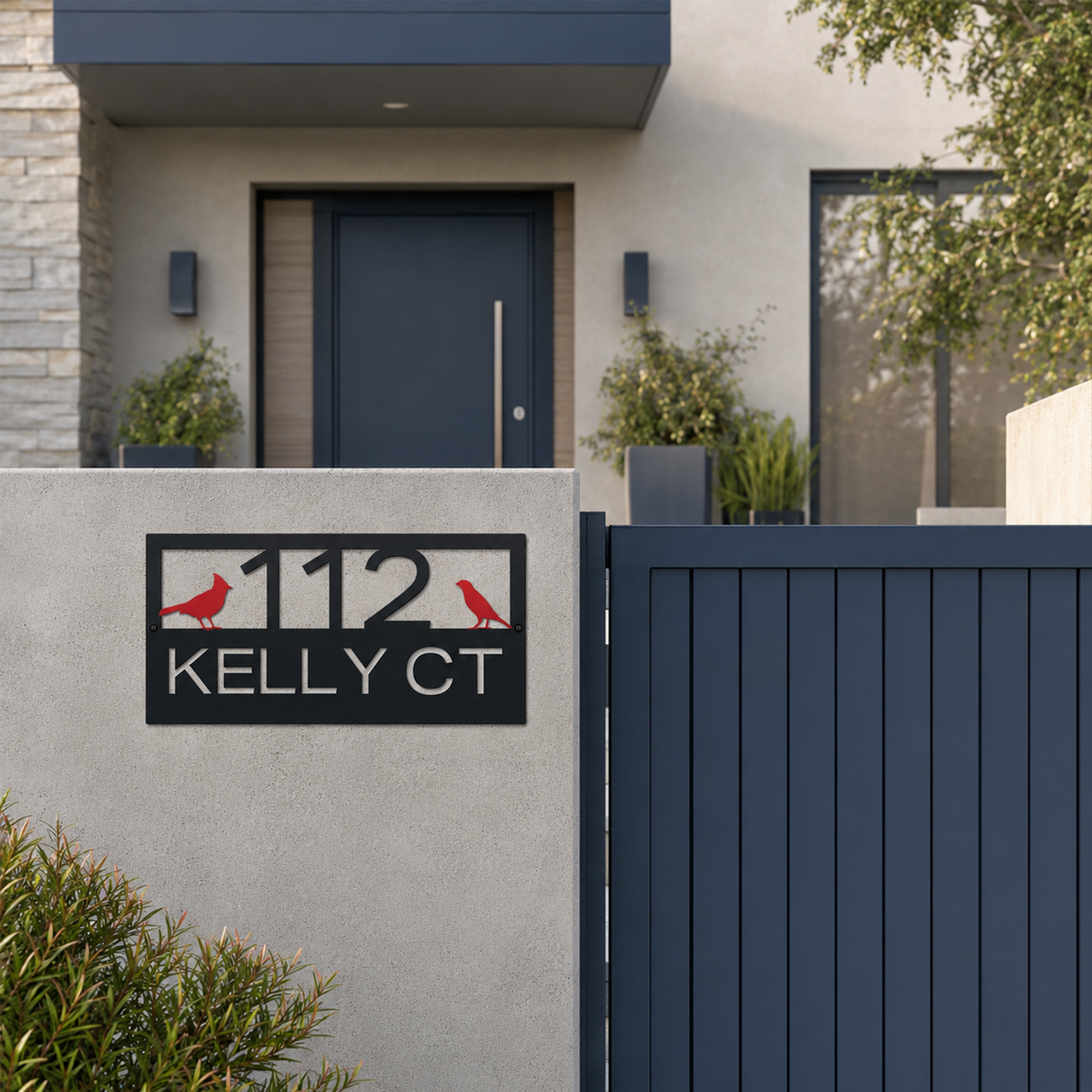 A matte black rectangular address plaque featuring "112 KELLY CT" and two red bird figures is mounted on a concrete wall alongside a dark blue vertical-slat gate. The upscale home entryway behind the gate shows a matching dark entry door, symmetrical potted plants, and modern stone cladding.