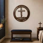 An espresso brown circular metal sign featuring a tree branch cross, the names "JOE & STEPHANIE," and the verse "JOHN 3:16" is mounted on a beige living room wall. Below the sign rests a dark wooden prayer bench with navy blue cushions, next to a small side table holding a freestanding wooden cross.