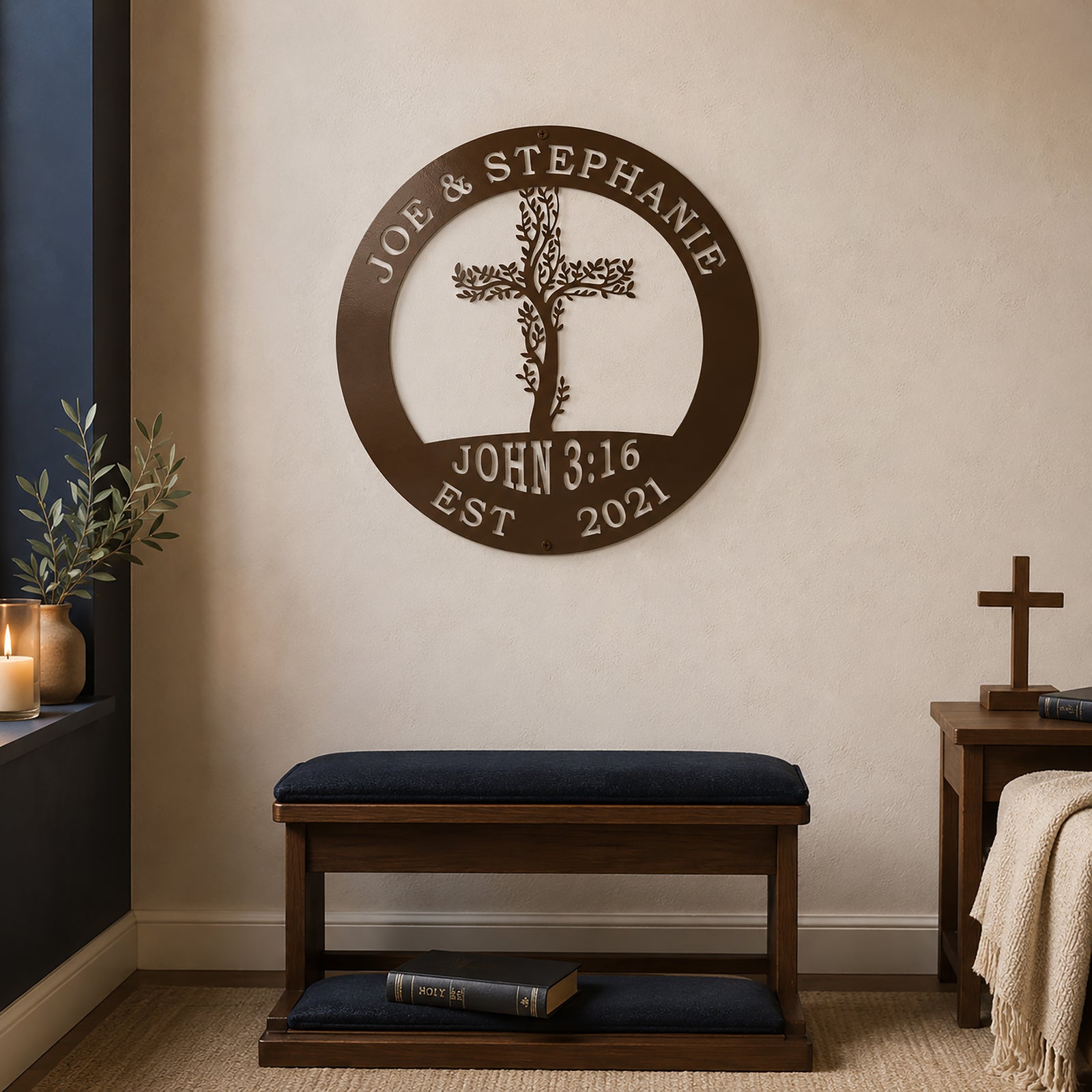An espresso brown circular metal sign featuring a tree branch cross, the names "JOE & STEPHANIE," and the verse "JOHN 3:16" is mounted on a beige living room wall. Below the sign rests a dark wooden prayer bench with navy blue cushions, next to a small side table holding a freestanding wooden cross.