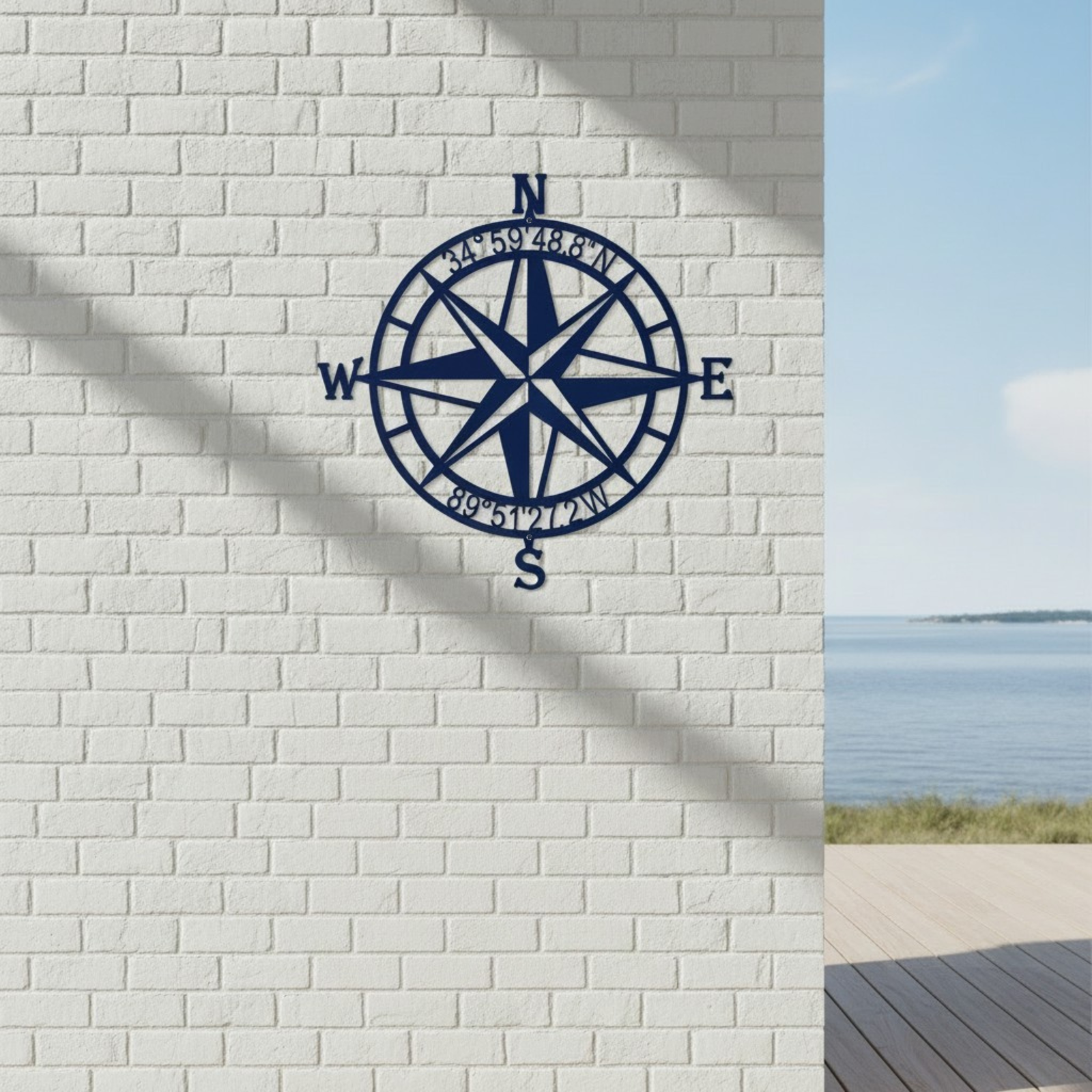 Custom compass rose metal sign — personalized coastal coordinates wall decor in navy blue powder-coated steel mounted on a white brick wall overlooking the waterfront.