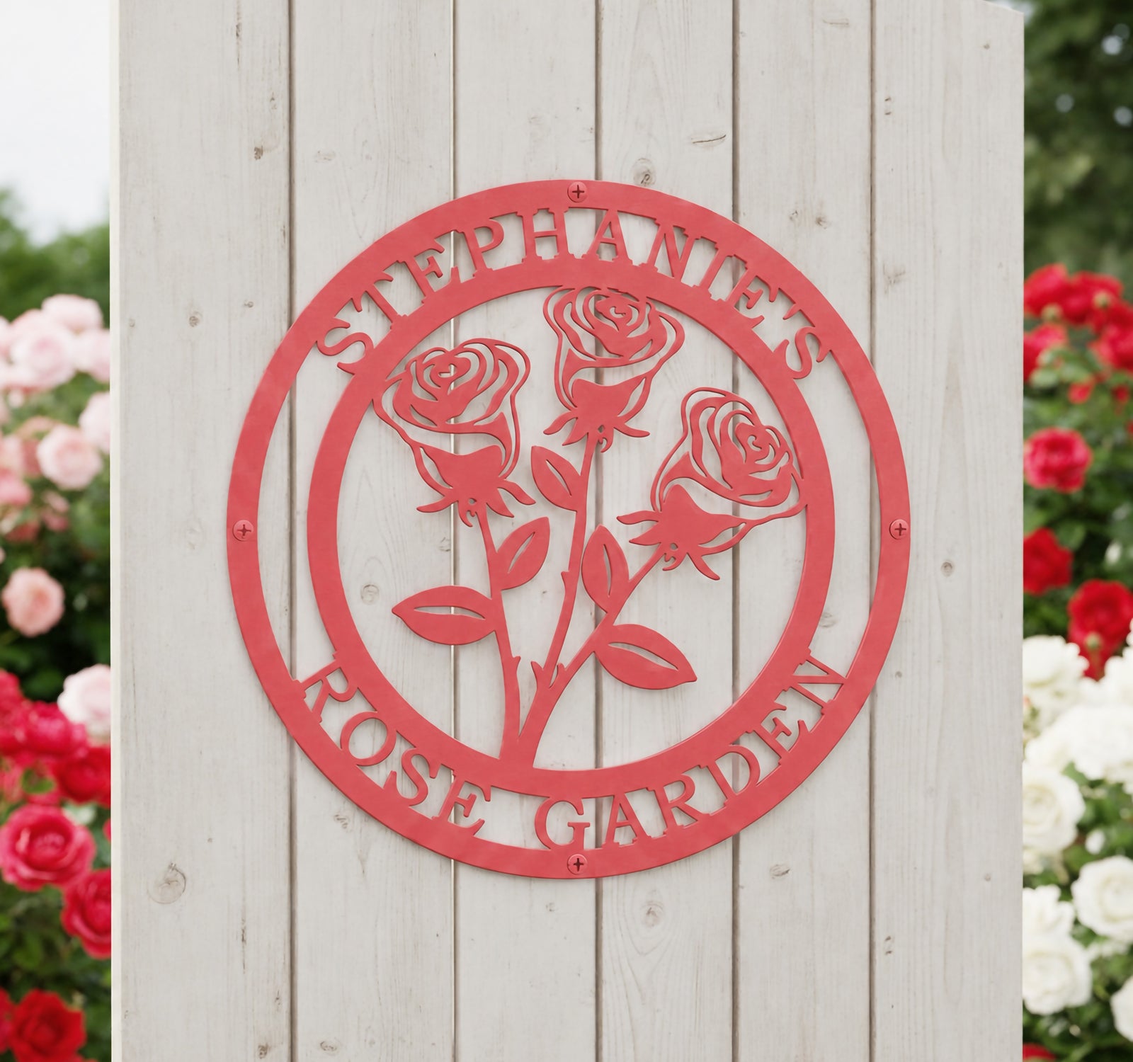 Matte coral pink personalized round metal garden plaque featuring a cutout three-rose design on a light gray vertical wood panel fence section surrounded by vibrant red bushes.