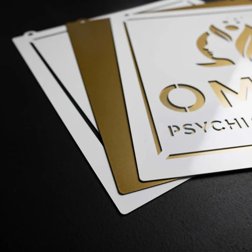 Close-up of white and gold signage made from cold-rolled American steel, featuring “OM,” partial “PSYCHI,” and stylized face logo.