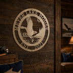 A circular custom metal family name plaque finished in a cream tone, featuring a laser-cut scene of an eagle catching a fish over a lake with the text "MEYER LAKE HOUSE EST. 2021". The piece is mounted to a rustic, dark-stained horizontal wood panel wall inside a cozy, dimly lit cabin room.