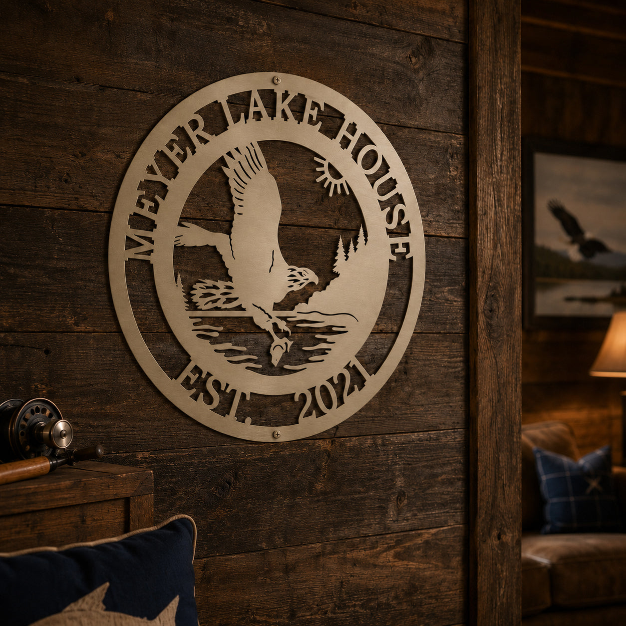 A circular custom metal family name plaque finished in a cream tone, featuring a laser-cut scene of an eagle catching a fish over a lake with the text "MEYER LAKE HOUSE EST. 2021". The piece is mounted to a rustic, dark-stained horizontal wood panel wall inside a cozy, dimly lit cabin room.