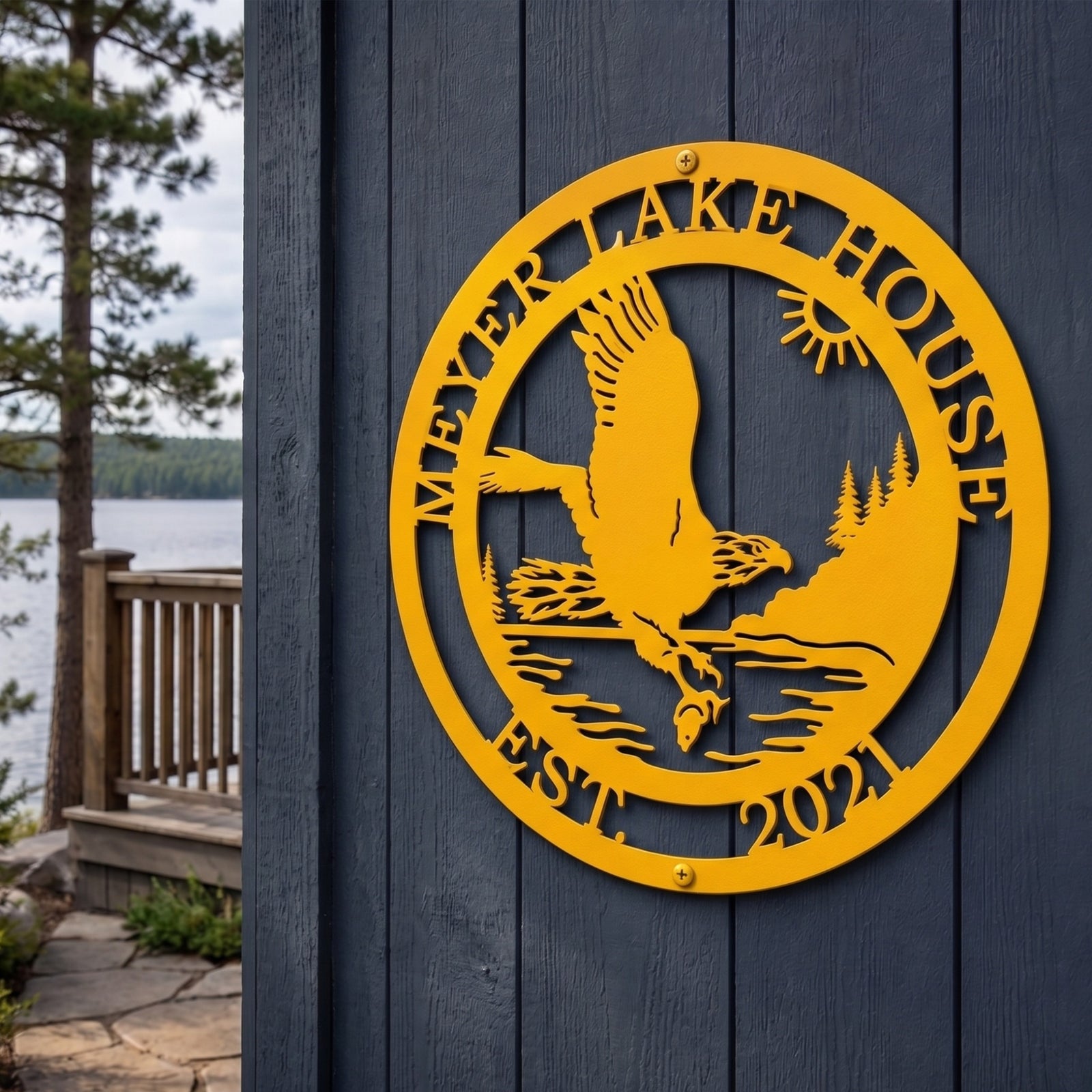 A round, laser-cut golden yellow metal sign mounted on dark blue wooden vertical siding. The central artwork shows an eagle catching a fish over water with a pine forest and sun background. The top text reads "MEYER LAKE HOUSE" and the bottom reads "EST. 2021". A lake and wooden deck railing are visible in the background.
