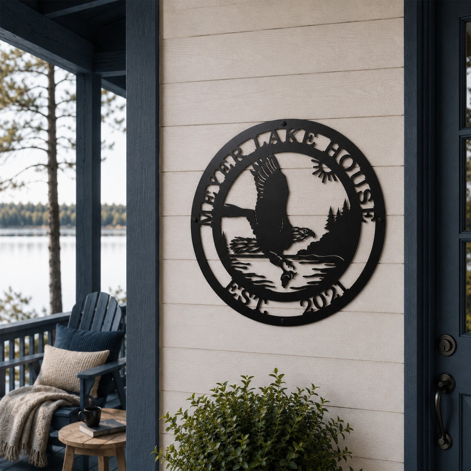 A round matte black laser-cut steel sign featuring a detailed eagle design, personalized with "MEYER LAKE HOUSE EST. 2021". The sign is mounted on light-colored horizontal siding on a covered lakeside deck with an Adirondack chair, side table, and green shrubbery visible.