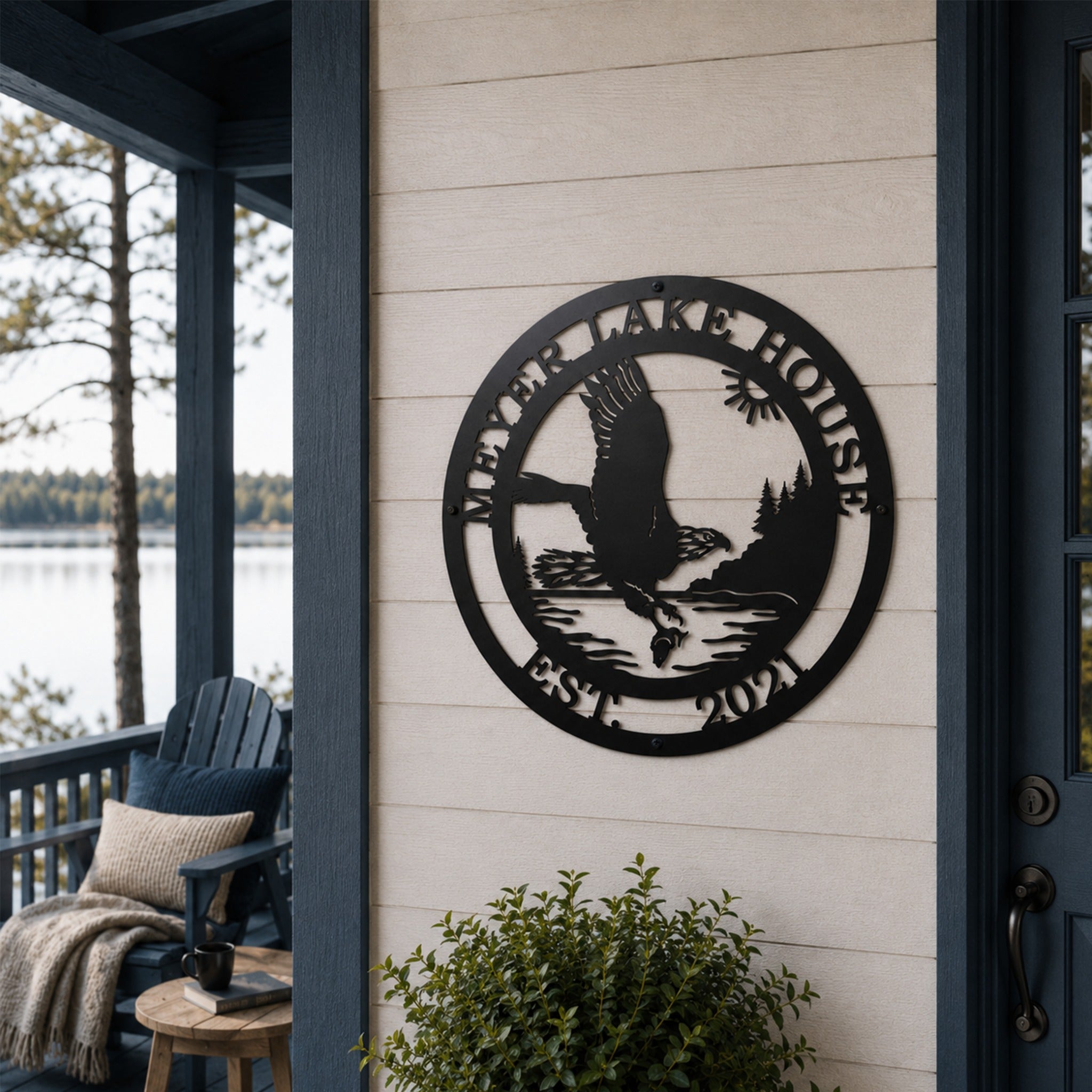 A round matte black laser-cut steel sign featuring a detailed eagle design, personalized with "MEYER LAKE HOUSE EST. 2021". The sign is mounted on light-colored horizontal siding on a covered lakeside deck with an Adirondack chair, side table, and green shrubbery visible.