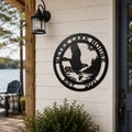 A circular matte black metal house sign displaying a scenic eagle design with the custom text "MEYER LAKE HOUSE EST. 2021". The sign is mounted onto off-white horizontal lap siding next to a dark-framed glass front door under an illuminated black outdoor lantern, with a blurry lakefront porch in the background.