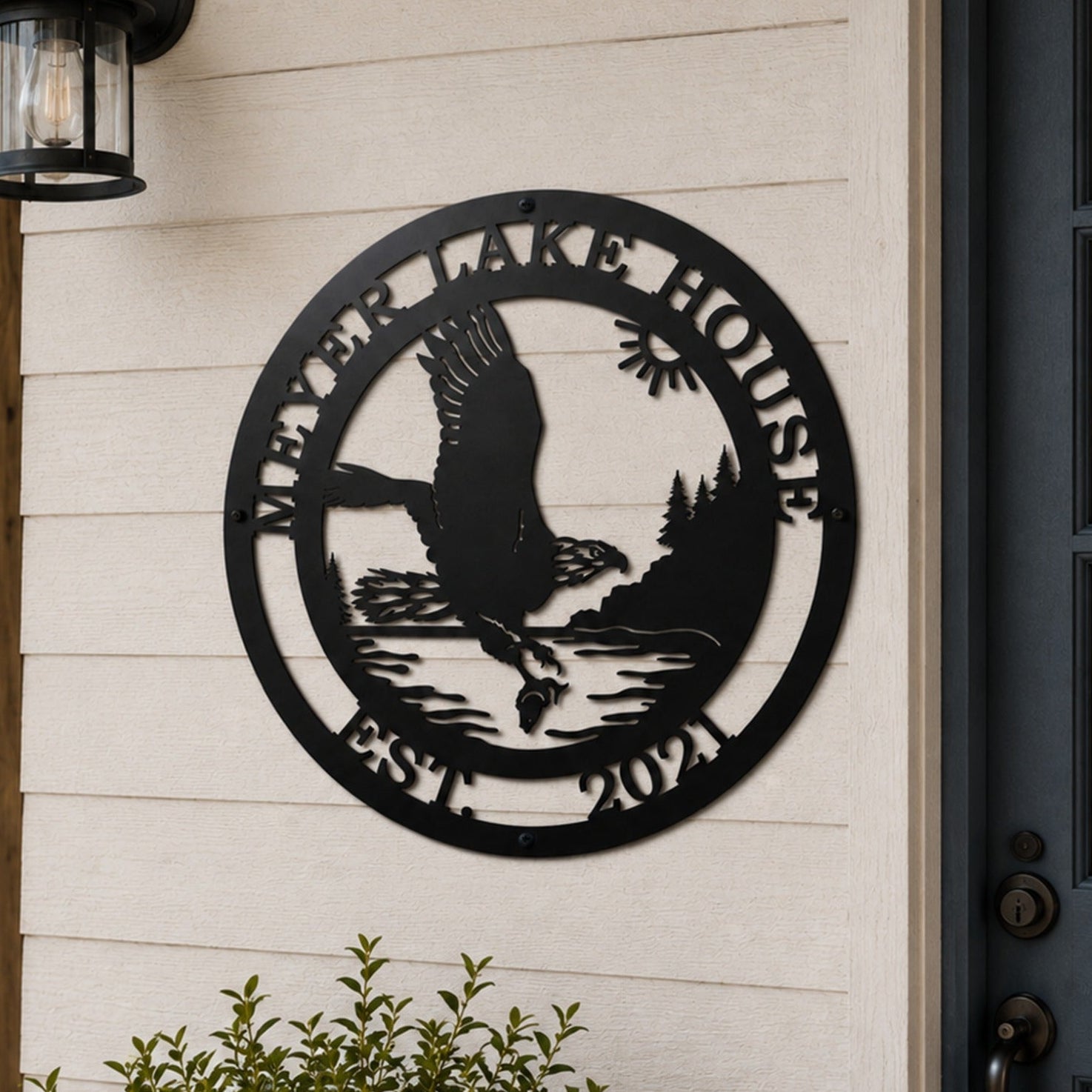 A circular matte black metal house sign displaying a scenic eagle design with the custom text "MEYER LAKE HOUSE EST. 2021". The sign is mounted onto off-white horizontal lap siding next to a dark-framed glass front door under an illuminated black outdoor lantern, with a blurry lakefront porch in the background.