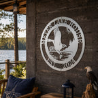 A round custom metal plaque in a sleek satin titanium finish, displaying an eagle catching a fish, personalized with "MEYER LAKE HOUSE EST. 2024". The sign is fixed to a textured dark brown accent wall on a rustic porch overlooking a lake, with an eagle figurine and small lantern placed on a wooden table below it.