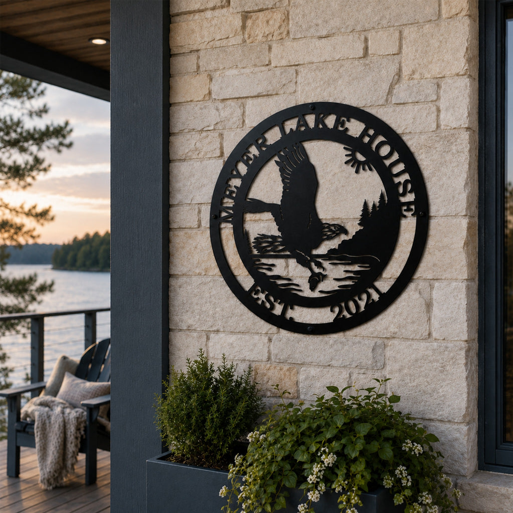 A round black metal sign featuring an eagle fishing scene bordered by the text "MEYER LAKE HOUSE EST. 2021". The plaque is anchored to a light beige stacked stone exterior column on a modern home patio overlooking a forest-lined lake during sunset.