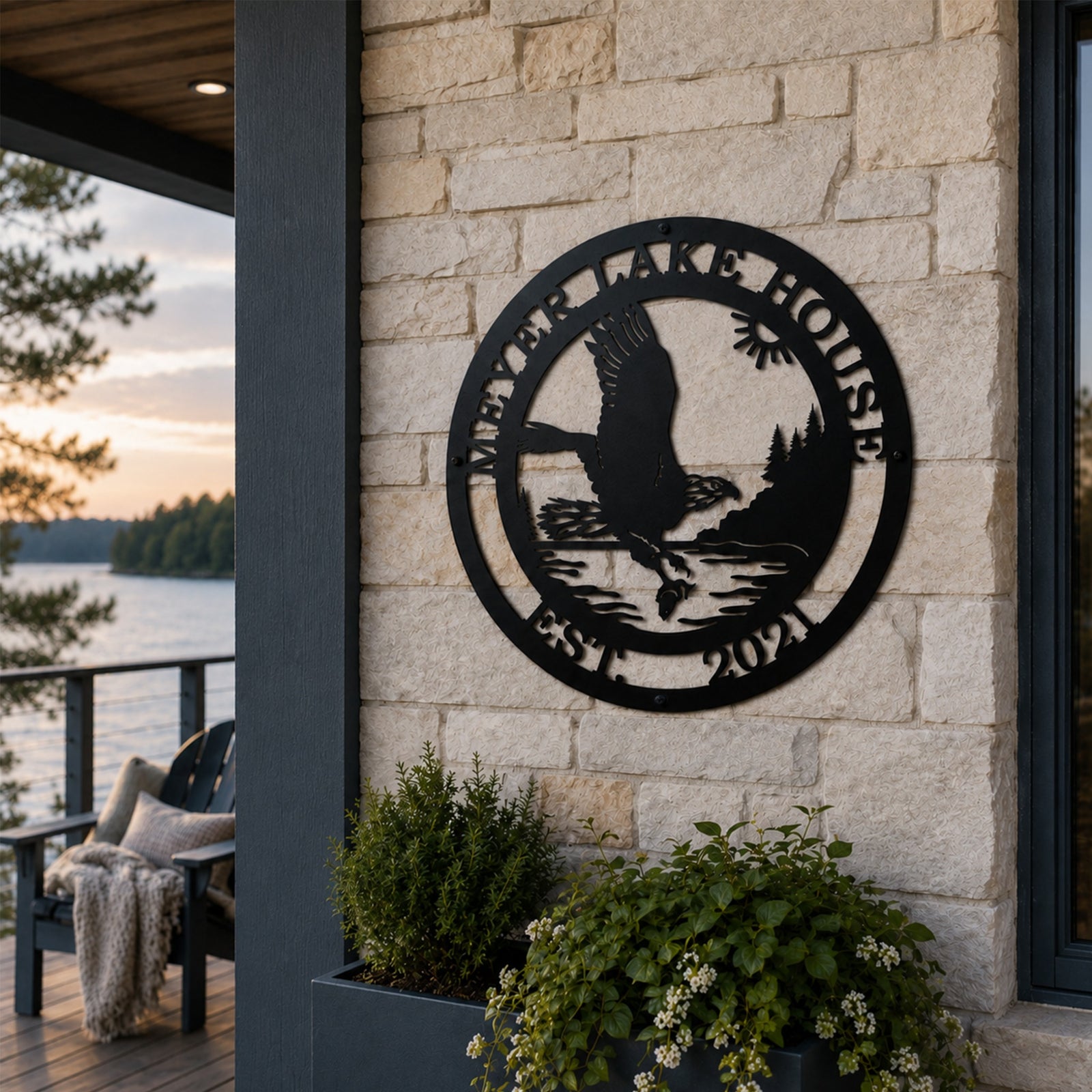 A round black metal sign featuring an eagle fishing scene bordered by the text "MEYER LAKE HOUSE EST. 2021". The plaque is anchored to a light beige stacked stone exterior column on a modern home patio overlooking a forest-lined lake during sunset.