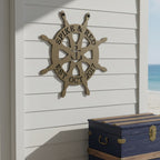 Gold finish of the custom circular ship wheel metal wall art styled on an exterior white siding wall above a rustic blue storage chest near the beach.