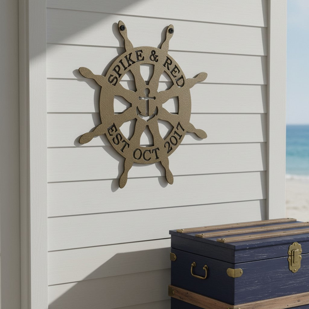 Gold finish of the custom circular ship wheel metal wall art styled on an exterior white siding wall above a rustic blue storage chest near the beach.