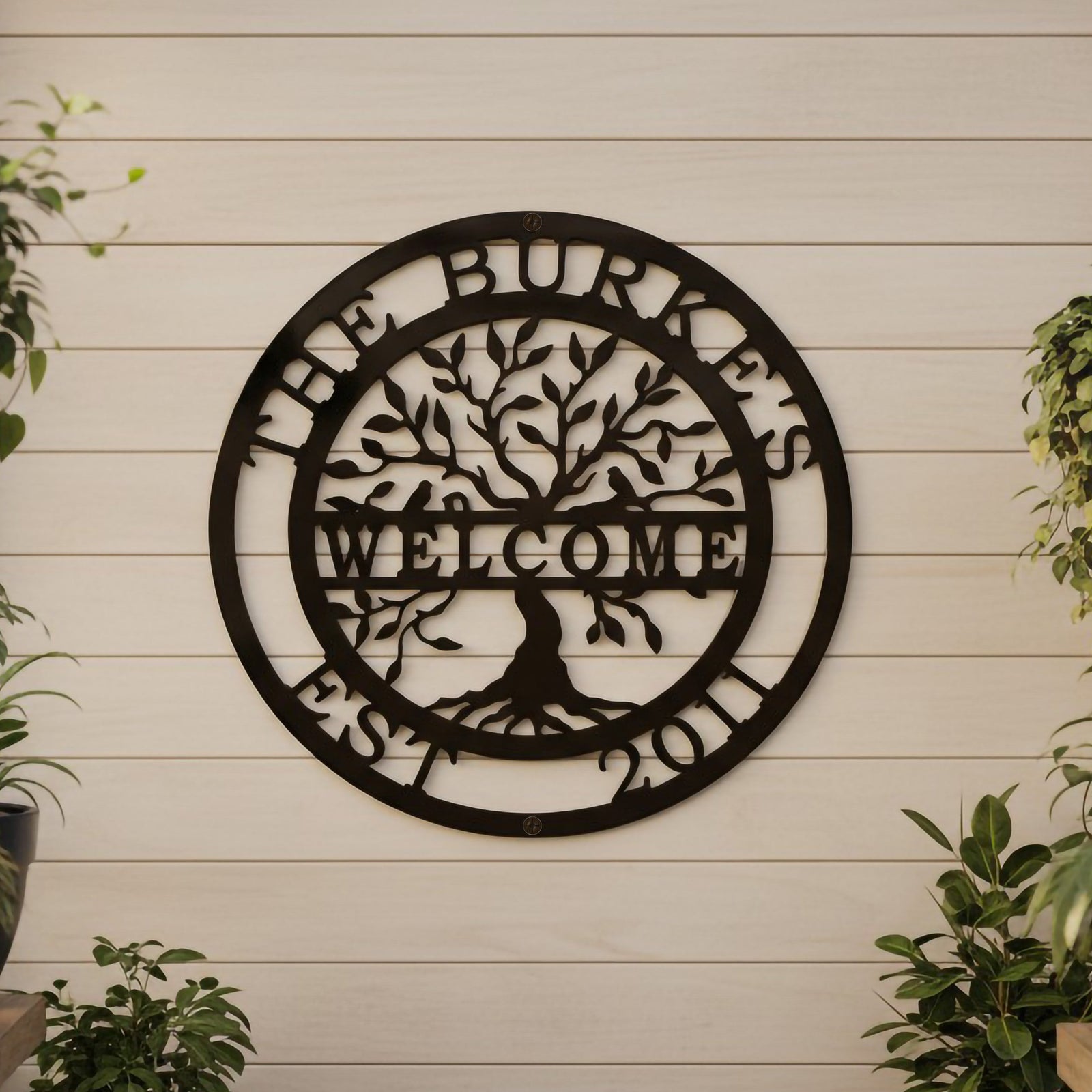 A dark brown circular metal tree of life sign reading "The Burkes Welcome Est. 2011," hanging on a light shiplap wall surrounded by hanging potted plants on wooden shelves.