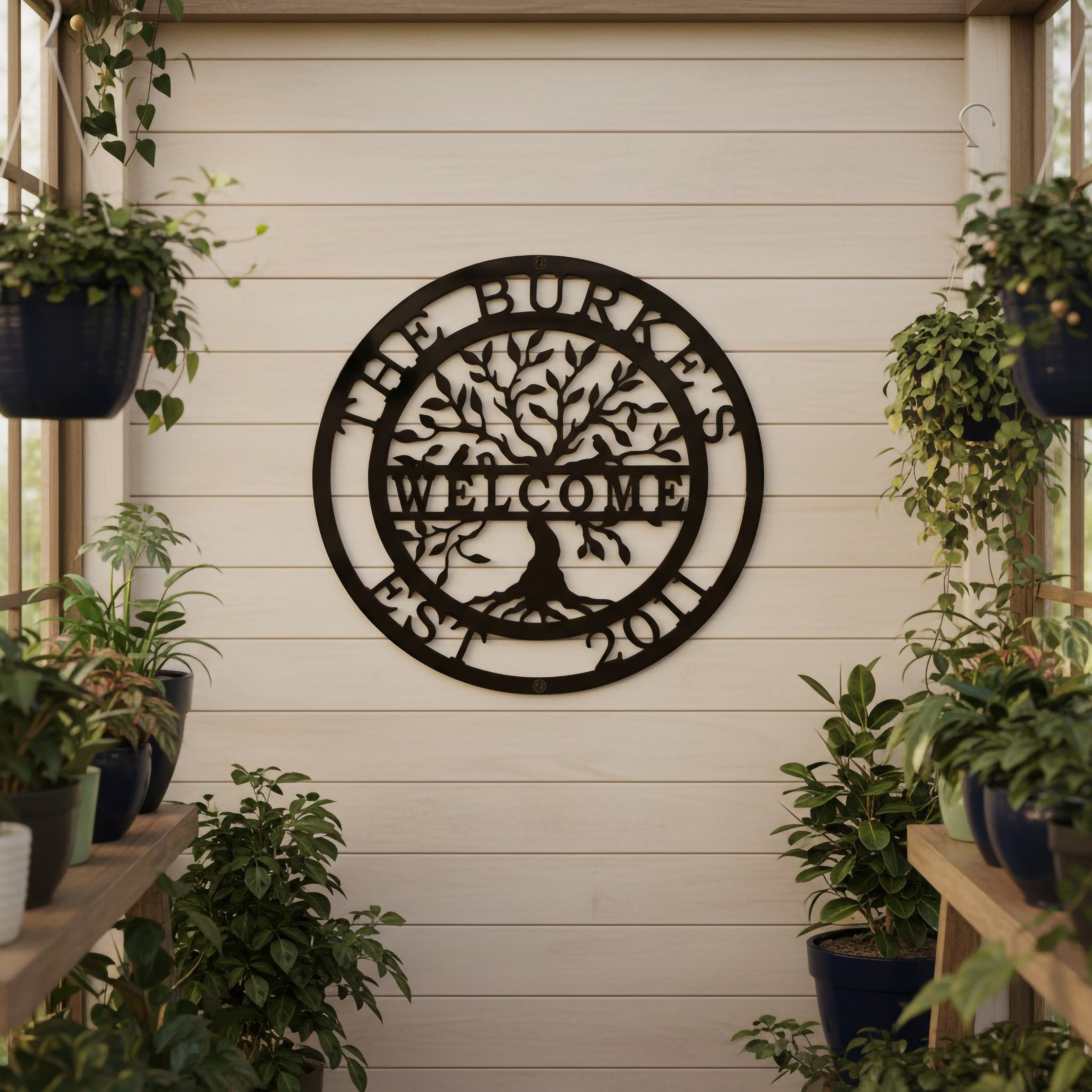 A dark brown circular metal tree of life sign reading "The Burkes Welcome Est. 2011," hanging on a light shiplap wall surrounded by hanging potted plants on wooden shelves.