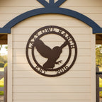 A round, laser-cut steel sign featuring a textured hammered bronze finish. The center design showcases the silhouette of an owl mid-flight with its wings spread, framed by a circular border containing the words "LAZY OWL RANCH" on top and "EST. 2016" along the bottom. The sign is mounted on a light cream horizontal lap siding accent wall under a decorative dark blue wooden archway.