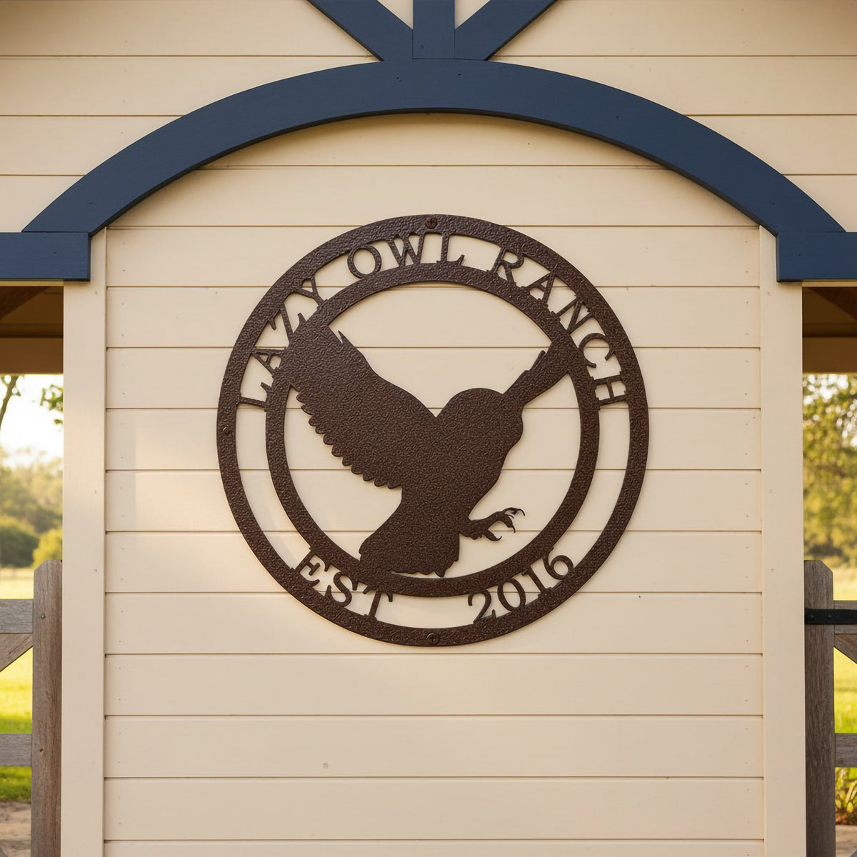 A round, laser-cut steel sign featuring a textured hammered bronze finish. The center design showcases the silhouette of an owl mid-flight with its wings spread, framed by a circular border containing the words "LAZY OWL RANCH" on top and "EST. 2016" along the bottom. The sign is mounted on a light cream horizontal lap siding accent wall under a decorative dark blue wooden archway.