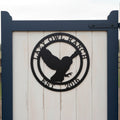A circular matte or satin black metal property sign depicting a flying owl silhouette, bordered by the custom inscriptions "LAZY OWL RANCH" and "EST. 2016". The sign is mounted on the white vertical wood paneling of a garden gate framed by deep navy blue posts.