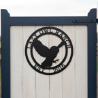 A circular matte or satin black metal property sign depicting a flying owl silhouette, bordered by the custom inscriptions "LAZY OWL RANCH" and "EST. 2016". The sign is mounted on the white vertical wood paneling of a garden gate framed by deep navy blue posts.