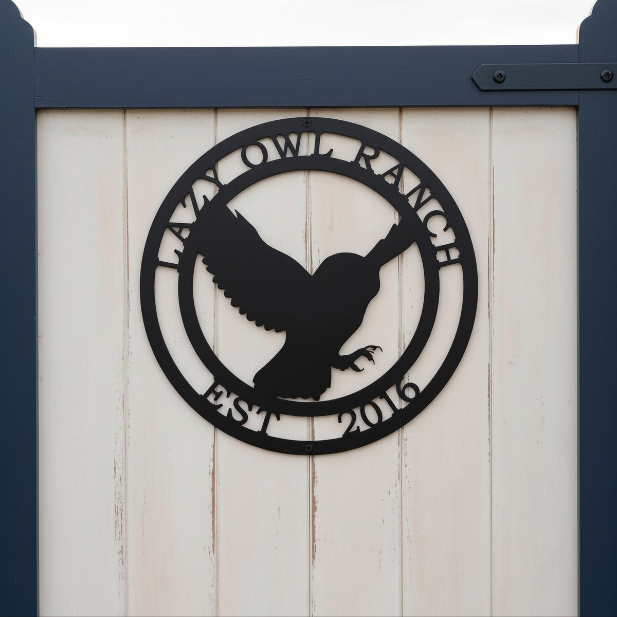 A circular matte or satin black metal property sign depicting a flying owl silhouette, bordered by the custom inscriptions "LAZY OWL RANCH" and "EST. 2016". The sign is mounted on the white vertical wood paneling of a garden gate framed by deep navy blue posts.