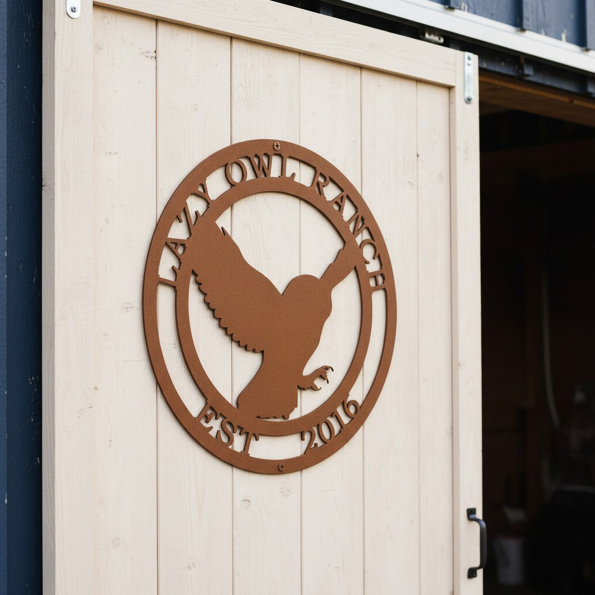 An angled view of a round custom metal plaque with an earthy, textured rust finish, showcasing an owl silhouette with outstretched wings. Personalized with the text "LAZY OWL RANCH EST. 2016", the sign is securely mounted to a light-colored vertical plank sliding barn door.