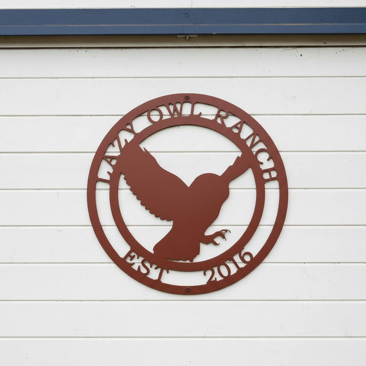 A circular custom metal sign in a smooth Tuscan copper finish, highlighting an owl in flight. The outer border is personalized with the laser-cut text "LAZY OWL RANCH" and "EST. 2016". The piece is installed directly on clean, white horizontal siding positioned beneath a dark blue trim piece.
