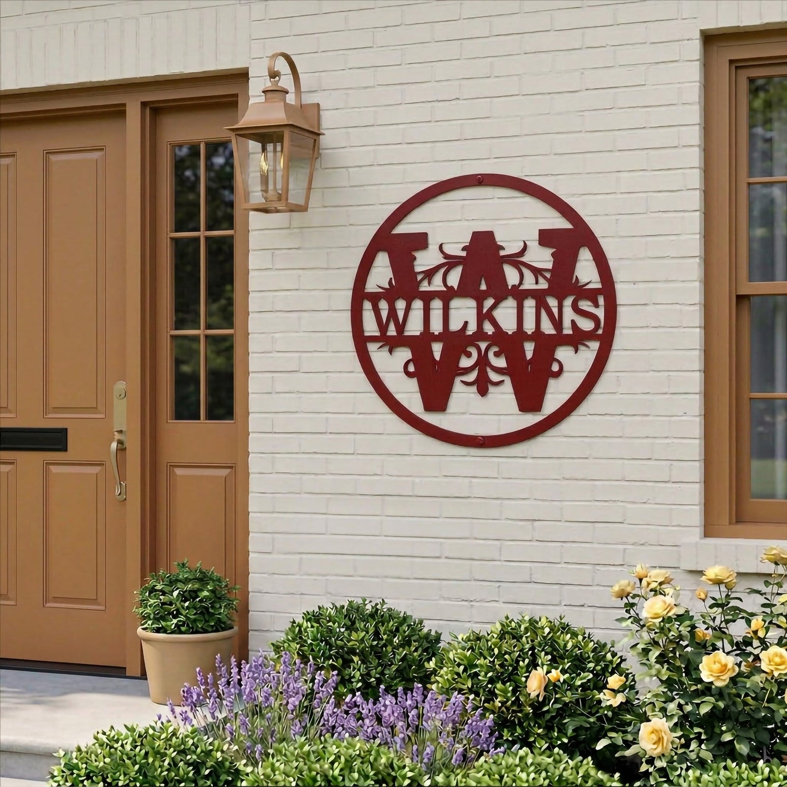 Rich garnet red customized round metal split monogram sign featuring the family name Wilkins, mounted on a white painted brick exterior wall beside a tan wooden front door with yellow flower beds below.
