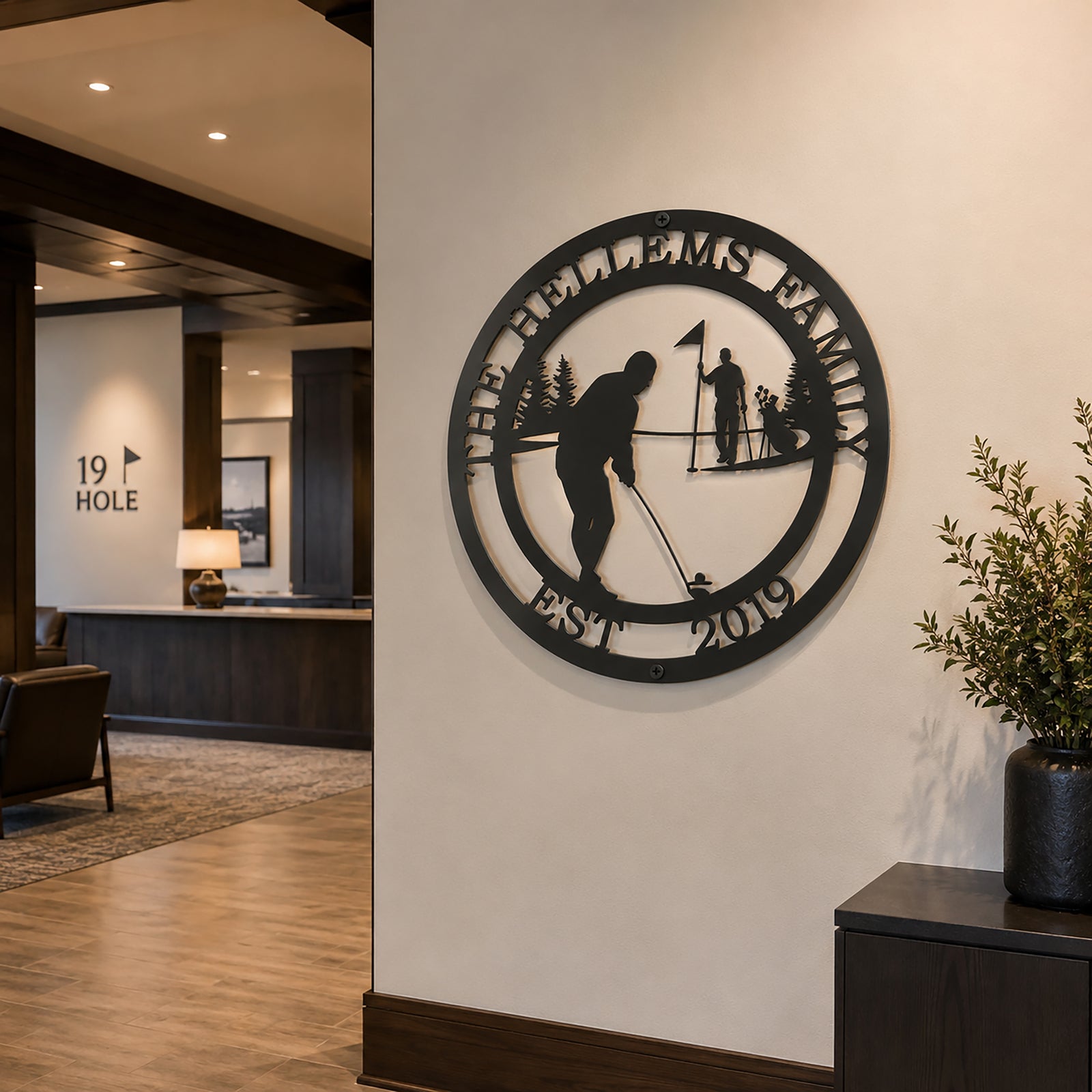 Matte black personalized golf silhouette metal sign with custom text The Hellems Family Est 2019 mounted on a white interior wall in a country club lobby next to a 19th Hole entryway.
