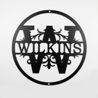 Flat view of a black circular personalized metal sign featuring a prominent letter W split monogram with elegant filigree accents and the custom surname Wilkins over a solid white background.