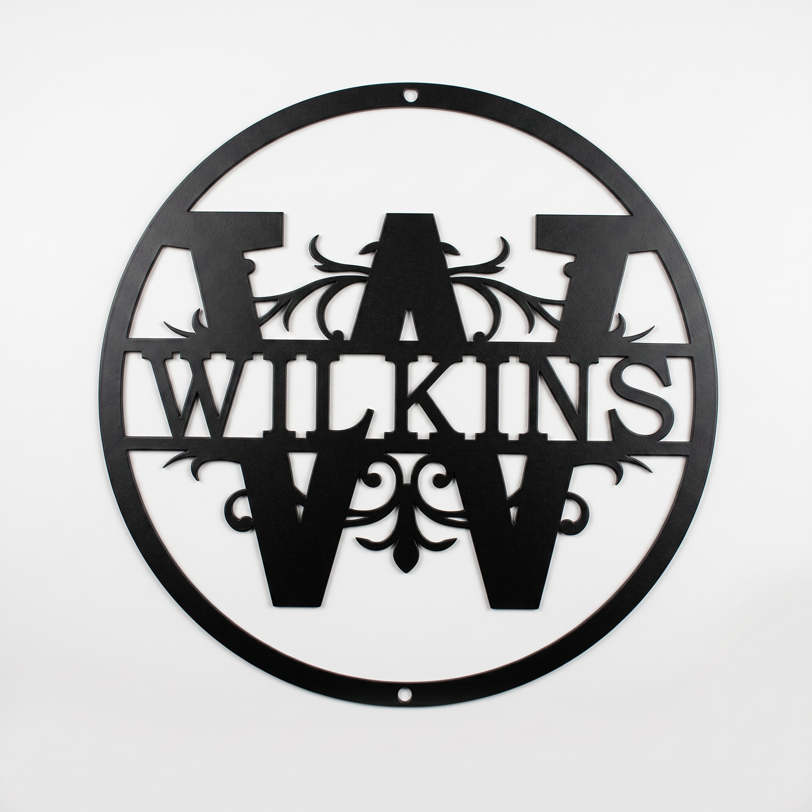 Flat view of a black circular personalized metal sign featuring a prominent letter W split monogram with elegant filigree accents and the custom surname Wilkins over a solid white background.