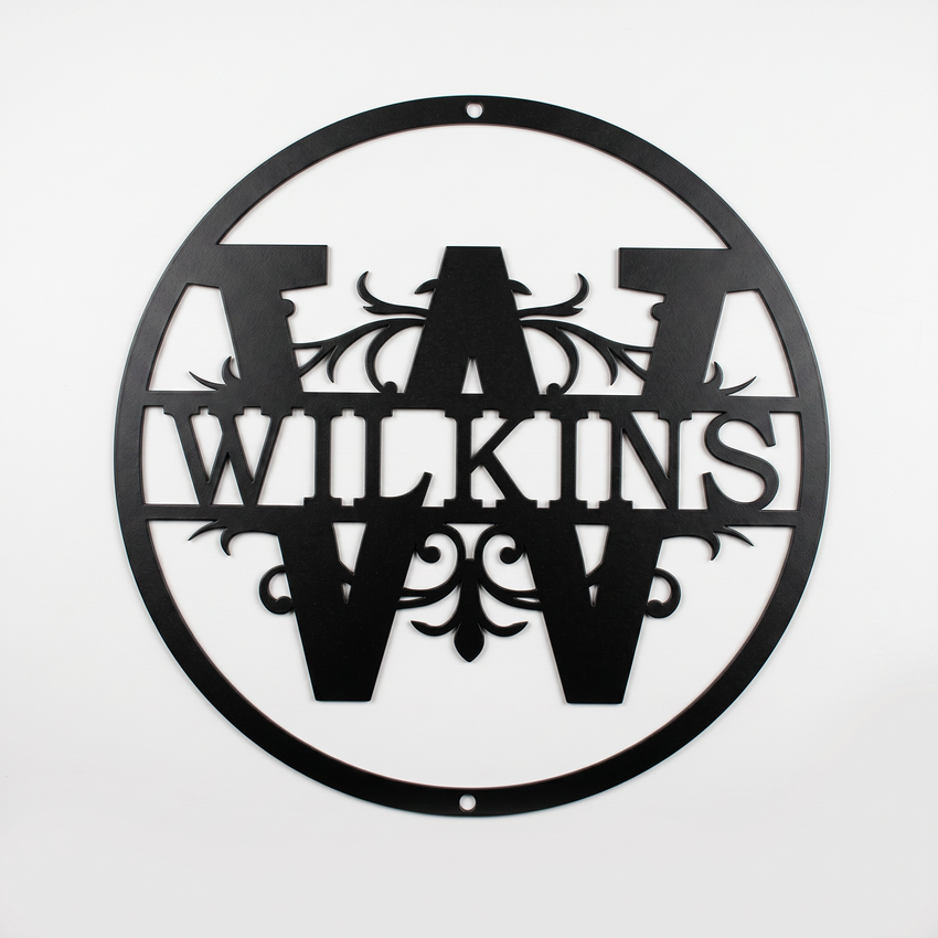 Flat view of a black circular personalized metal sign featuring a prominent letter W split monogram with elegant filigree accents and the custom surname Wilkins over a solid white background.