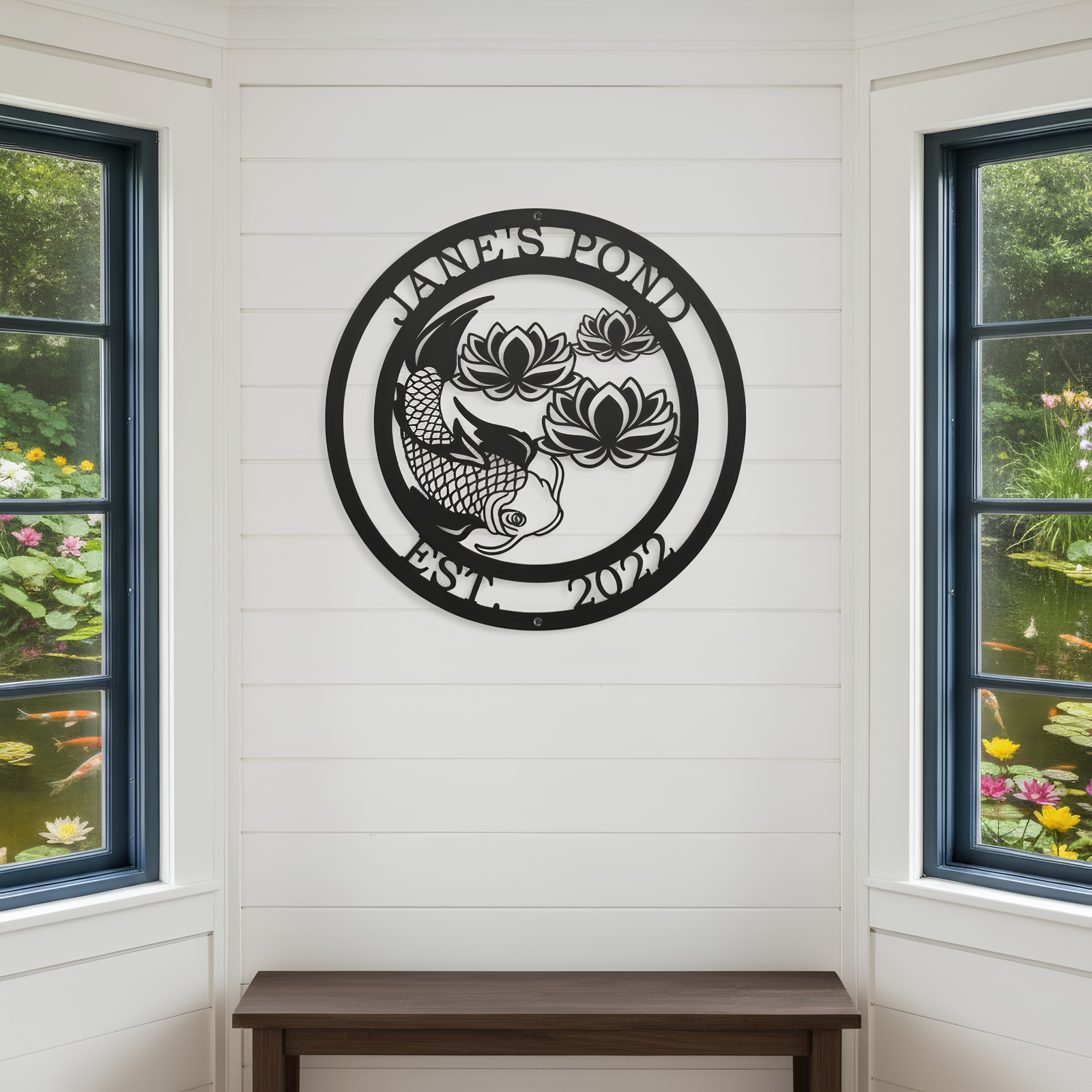 A circular black metal sign reading "Jane's Pond Est. 2022" with a detailed koi fish and lotus flowers, mounted indoors on a white shiplap wall between two large windows.