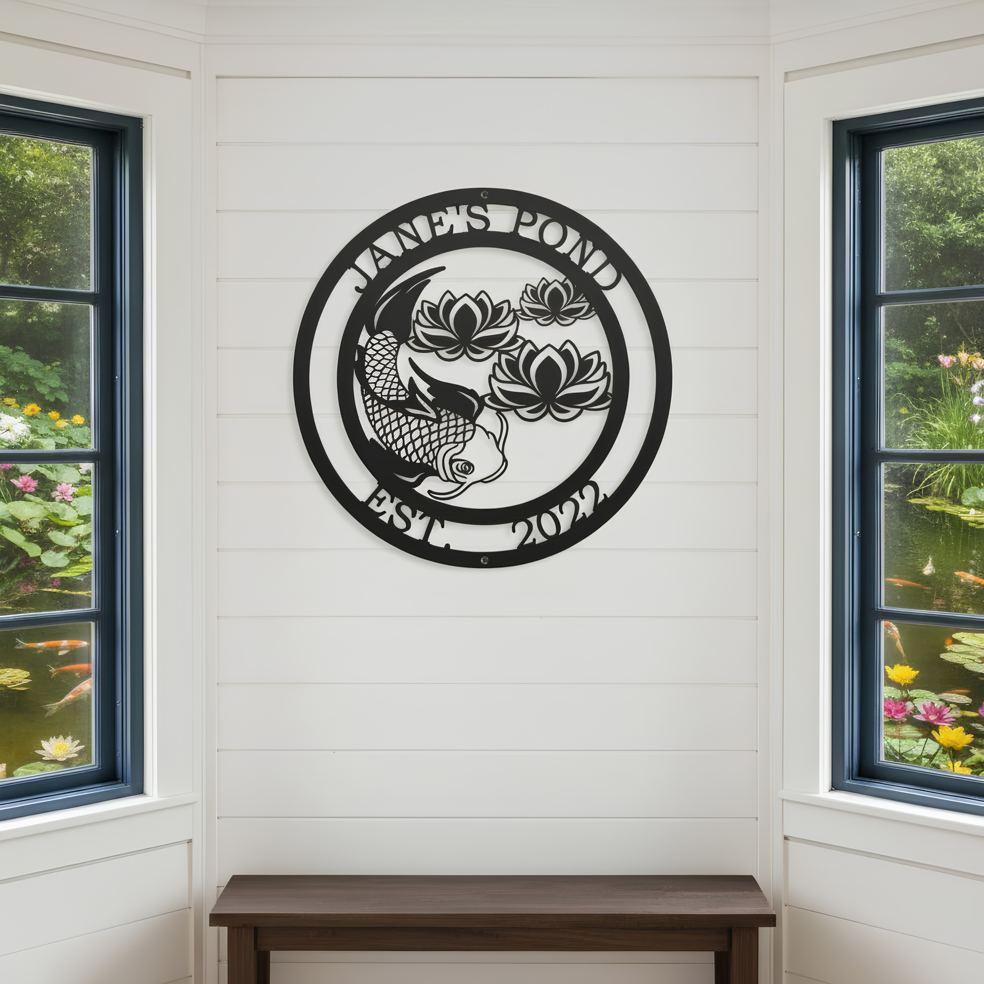 A circular black metal sign reading "Jane's Pond Est. 2022" with a detailed koi fish and lotus flowers, mounted indoors on a white shiplap wall between two large windows.