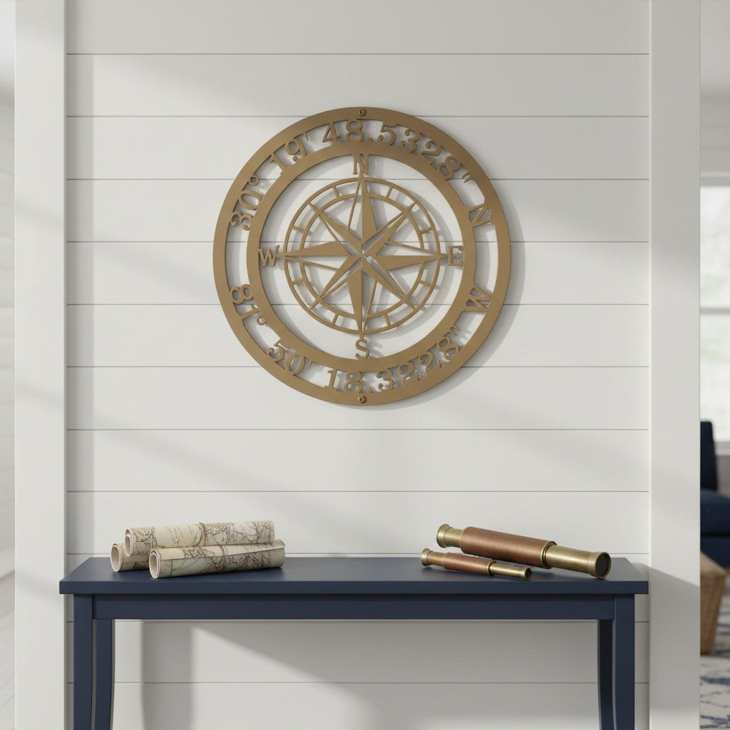 A gold circular metal compass sign customized with geographic coordinates along the outer border, mounted on a white shiplap wall above a dark blue console table decorated with rolled maps and a vintage brass telescope.
