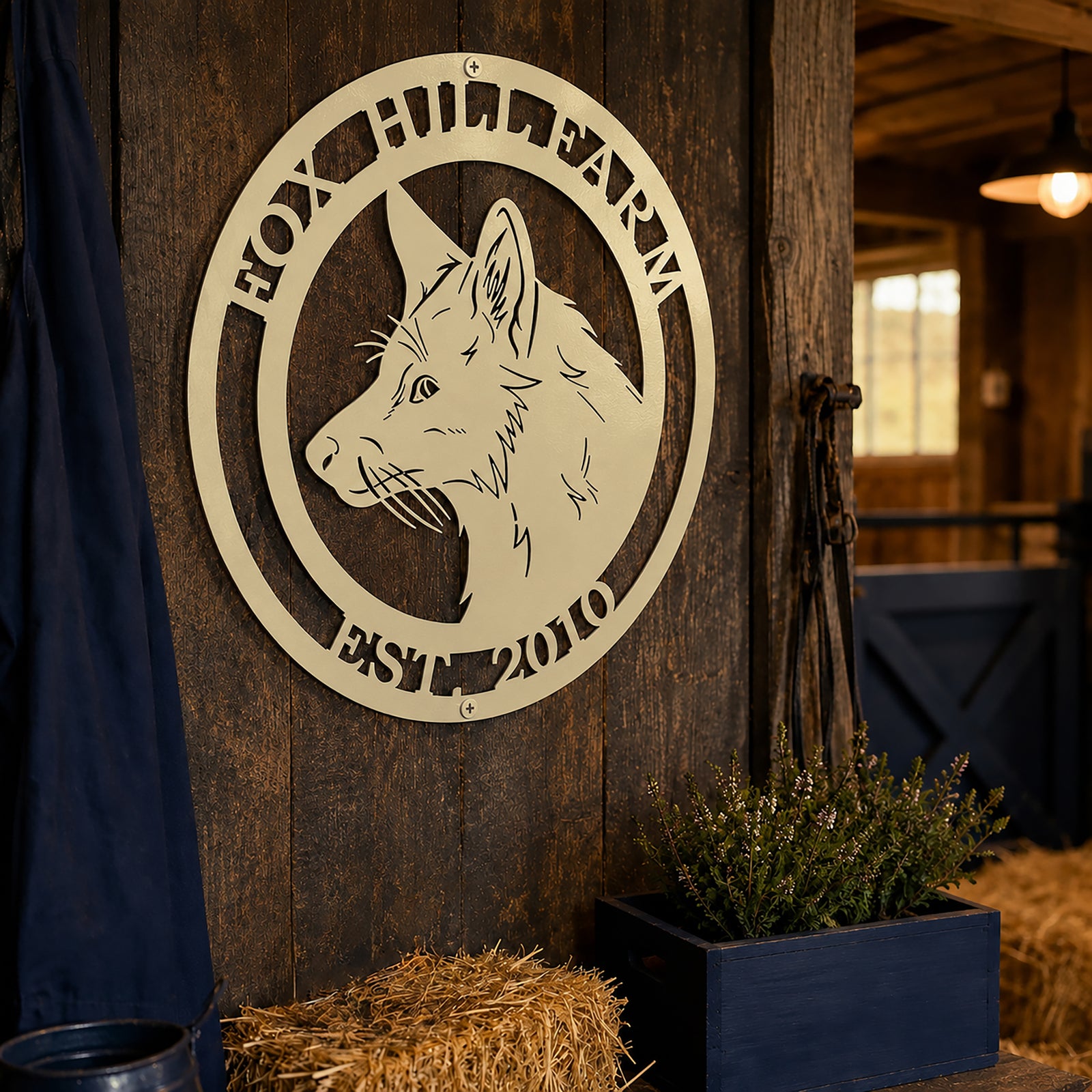 An ivory-colored circular metal sign featuring a fox head silhouette mounted on a dark, rustic wood paneled wall. The top of the sign says "FOX HILL FARM" and the bottom says "EST. 2010", positioned next to a small wooden crate of plants and a bundle of straw inside a barn.
