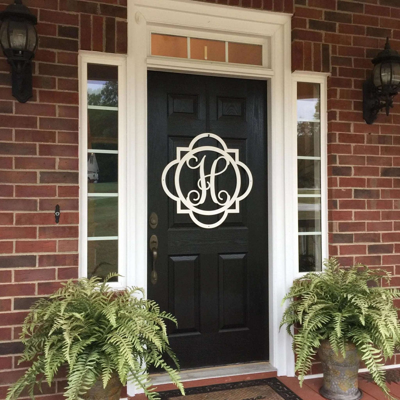 Custom metal monogram front door sign in white displayed on black door with brick exterior and porch plants