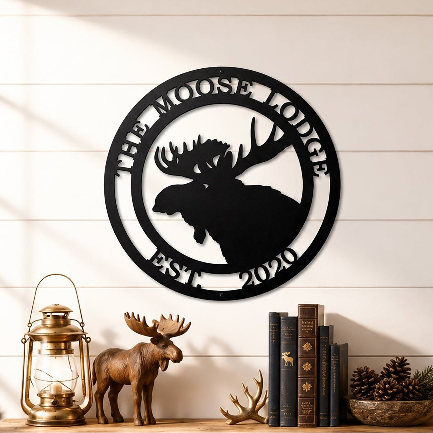 Black custom moose cabin metal wall sign displayed above wooden shelf with lantern books pinecones and lodge décor