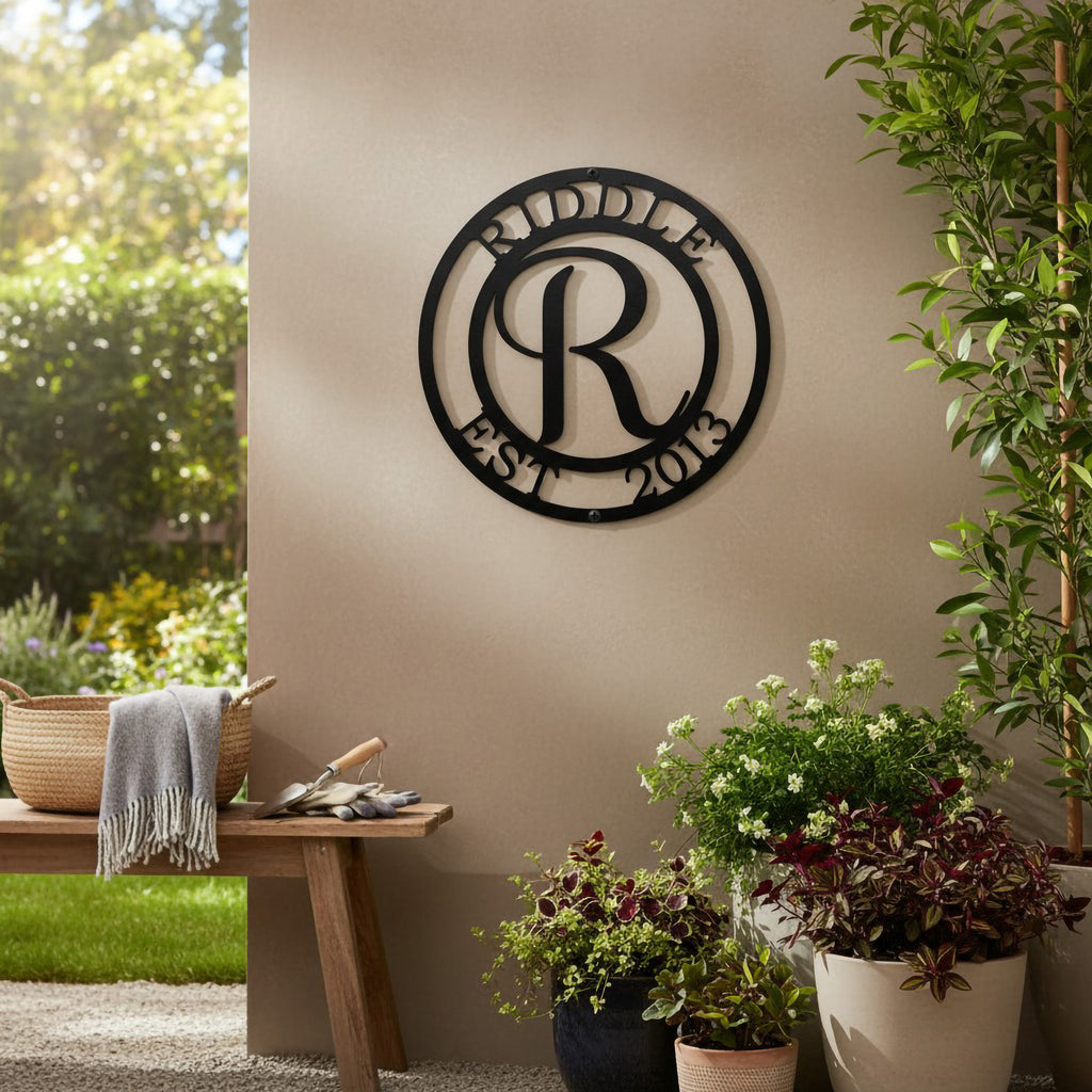 Matte black personalized letter R initial family metal sign mounted on a tan exterior porch wall above patio planters, potted greenery, and a wooden bench with a throw blanket.